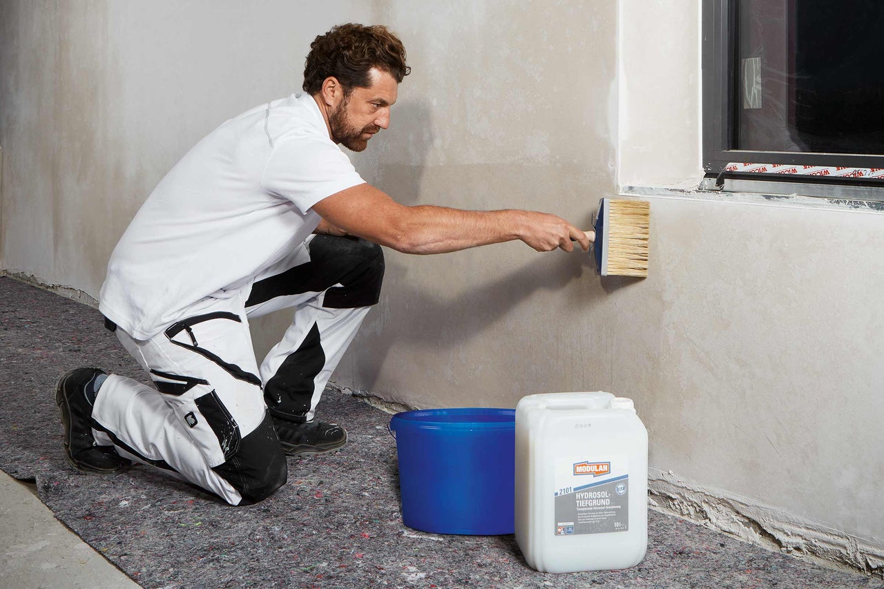 Un bărbat aplică grund pe un perete cu o bidinea lată, lângă el se află o găleată și un bidon cu grund de profunzime.
