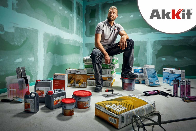 Scenă cu diverse produse Akkit, inclusiv materiale de umplutură, chituri și adezivi, în fața unui șantier cu pereți tencuiți. Un bărbat stă așezat pe un teanc de saci Akkit. Logo Akkit.