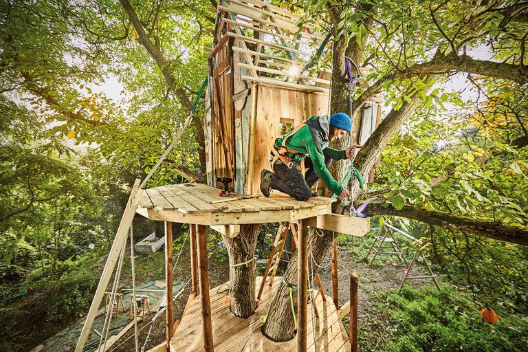 Un bărbat construiește o casă în copac cu structură din lemn în grădină.