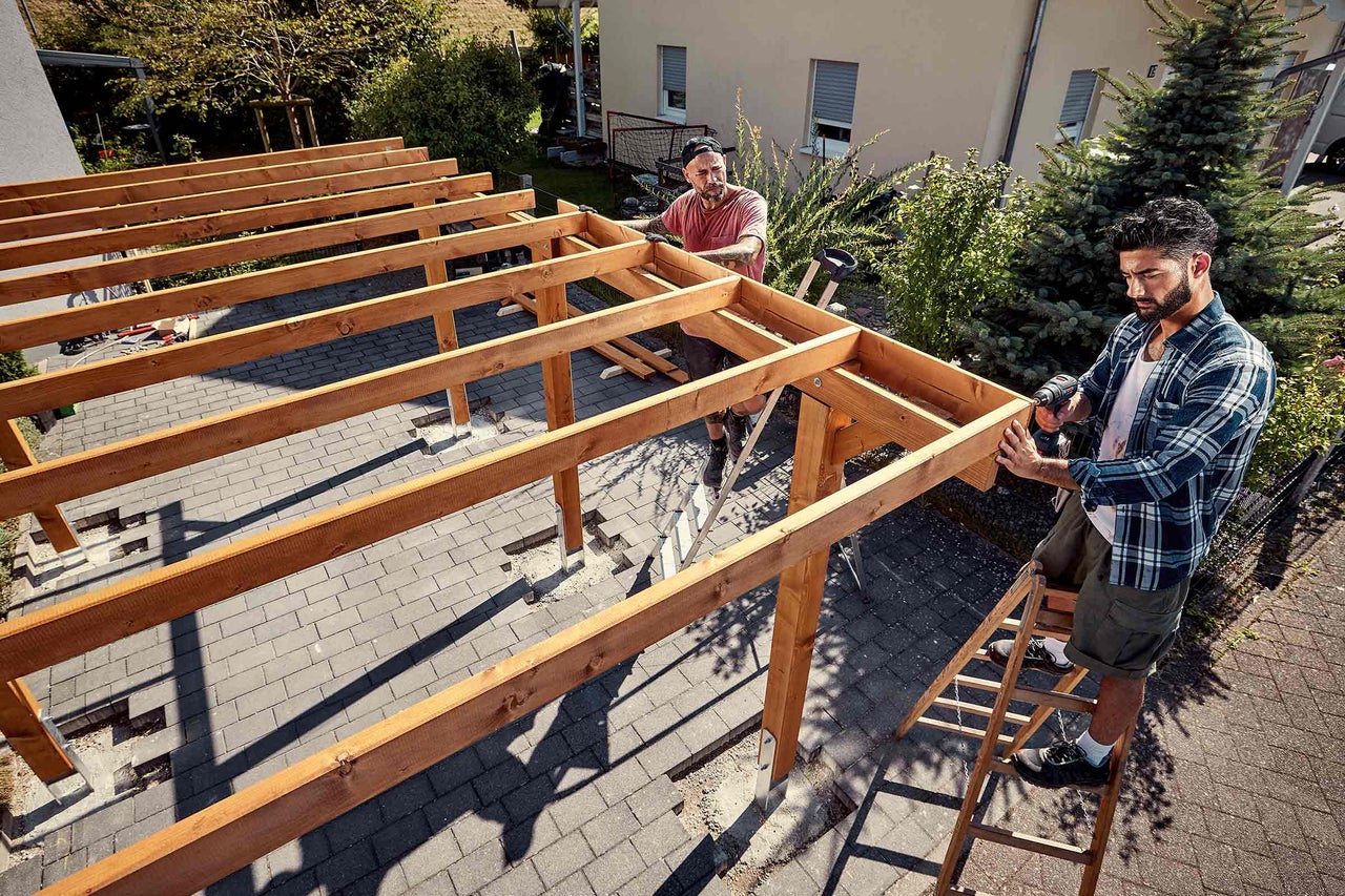 Doi meșteri montează o structură din lemn pentru o copertină de terasă folosind o mașină de găurit cu acumulator pe o terasă pavată din grădină.