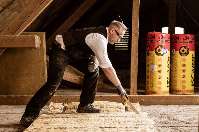 Un bărbat taie material izolant într-un pod. În fundal se află role cu material izolant.