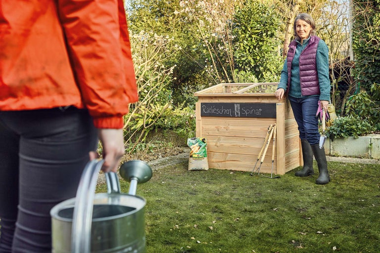 Scenă de grădină cu jardinieră înălțată, stropitoare, femeie și unelte de grădină în grădină