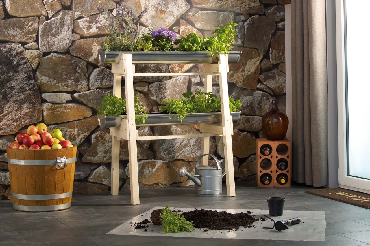 Jardinieră înaltă din lemn cu ierburi și legume, găleată cu mere, stropitoare și unelte de grădinărit în fața unui zid de piatră