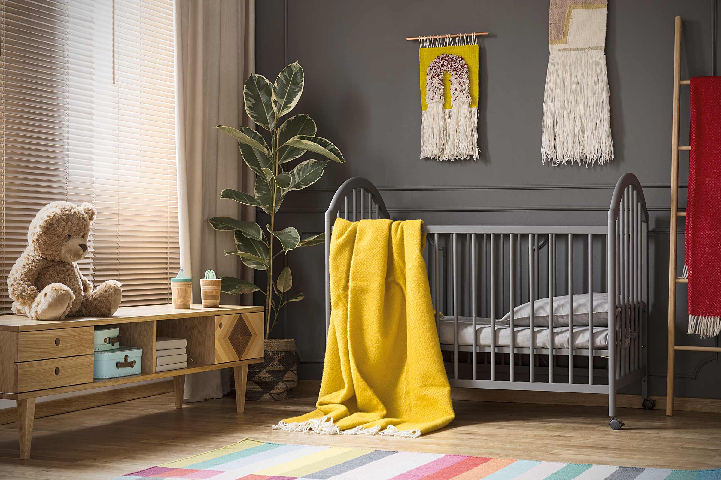 Cameră de copii cu pătuț, comodă, plantă de interior și covoare decorative pentru o atmosferă confortabilă.