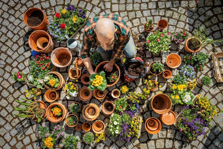 Bărbat plantând flori în ghivece afară, înconjurat de pământ, stropitoare și unelte de grădină.
