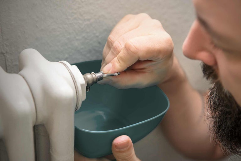 Bărbat aerisind caloriferul cu o cheie de aerisire și un recipient