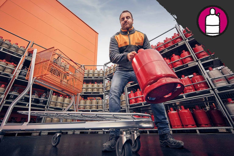 Bărbat care umple un cărucior de cumpărături cu o butelie de gaz în fața unui raft cu butelii de gaz