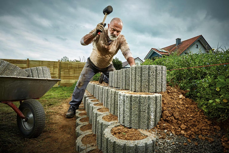 Un bărbat construiește un zid de grădină din pietre de beton cu un ciocan de cauciuc, roabă în fundal.