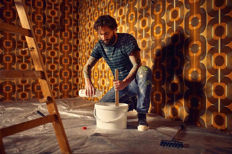 Bărbat amestecând vopsea într-o găleată pentru renovare într-o cameră cu tapet retro.