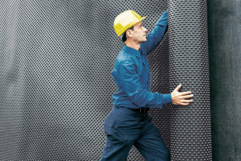 Un bărbat cu cască de protecție montează folie cu alveole pentru etanșarea construcțiilor.