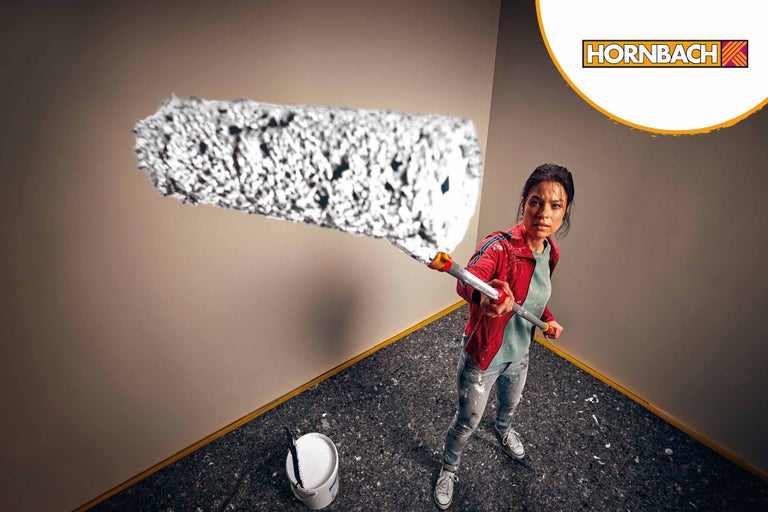 Femeie vopsește un perete cu trafaletul, găleată cu vopsea în cameră, logo Hornbach