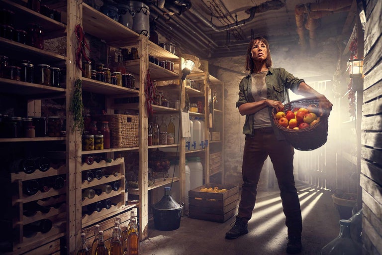 O femeie stă într-o pivniță cu rafturi pline de conserve, sticle și provizii și ține un coș cu mere.