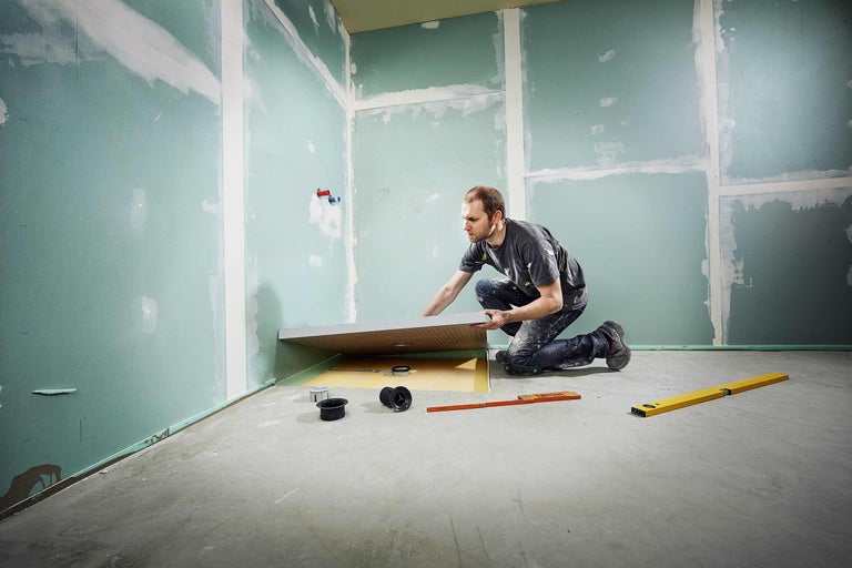 Un bărbat instalează o cadă de duș într-o baie cu pereți din gips-carton și instrumente de nivelare.