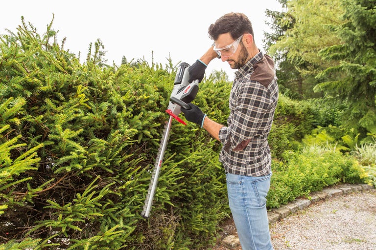 Un bărbat taie un gard viu cu o foarfecă de gard viu fără fir pentru grădinărit.