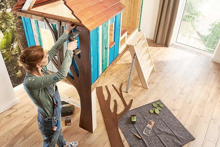 Femeie asamblând o căsuță de joacă pentru copii din lemn cu o mașină de găurit, cu scară și unelte