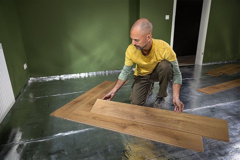 Un bărbat montează parchet laminat pe un strat izolator într-o cameră.