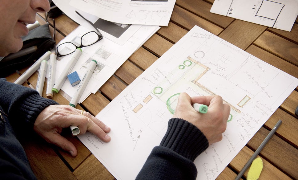 Un bărbat proiectează un plan de grădină cu markere pe o masă de lemn, reprezentând diverse elemente de horticultură.