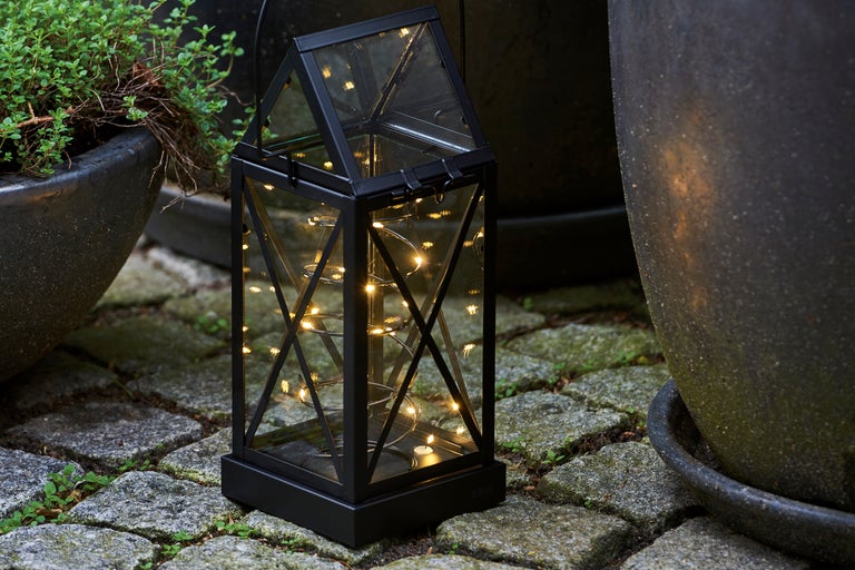 Lanternă decorativă de grădină cu șir de lumini lângă jardiniere pe pavaj