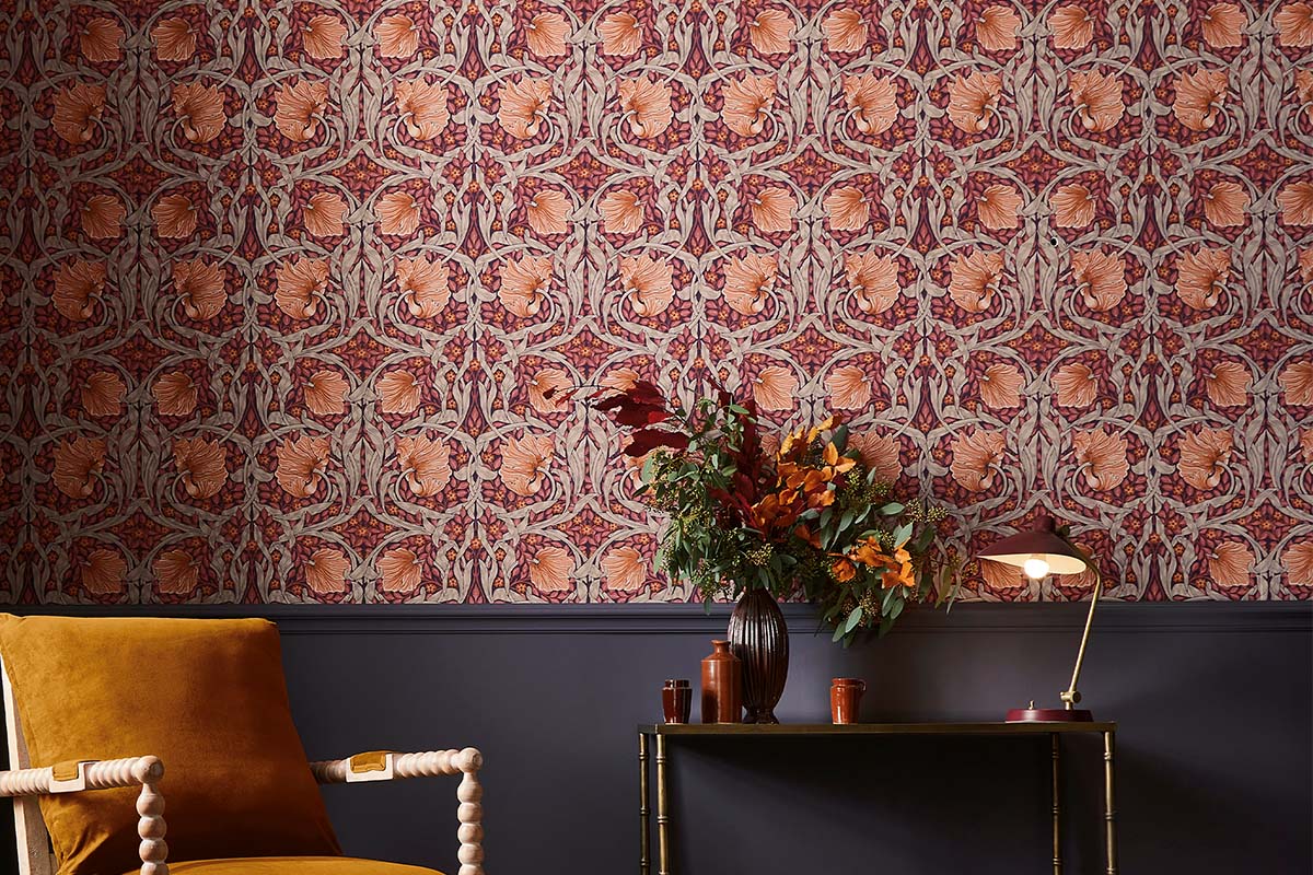 Cameră decorativă cu tapet cu model floral, fotoliu, masă cu vază de flori și lampă de masă.