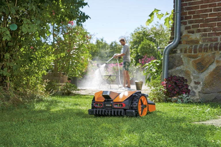 Robot de tuns iarba pe un gazon, cu o persoană care curăță mobilierul de grădină în fundal