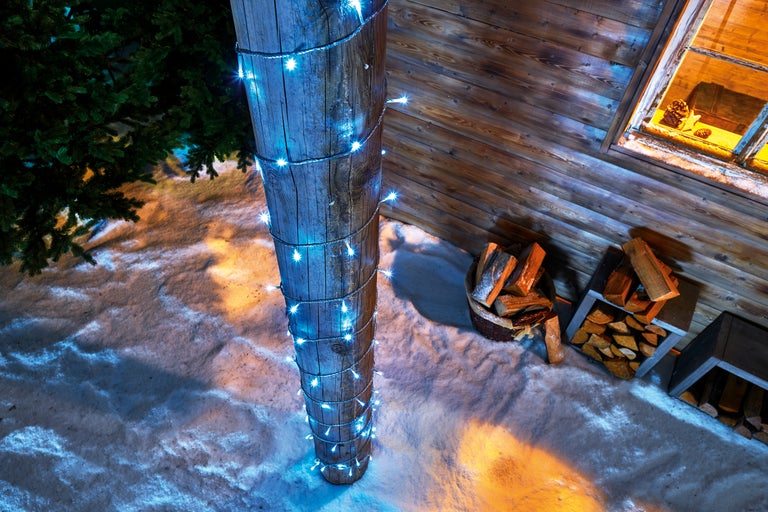 Stâlp de lemn cu șir de lumini, casă de lemn, lemne de foc în grădina de iarnă
