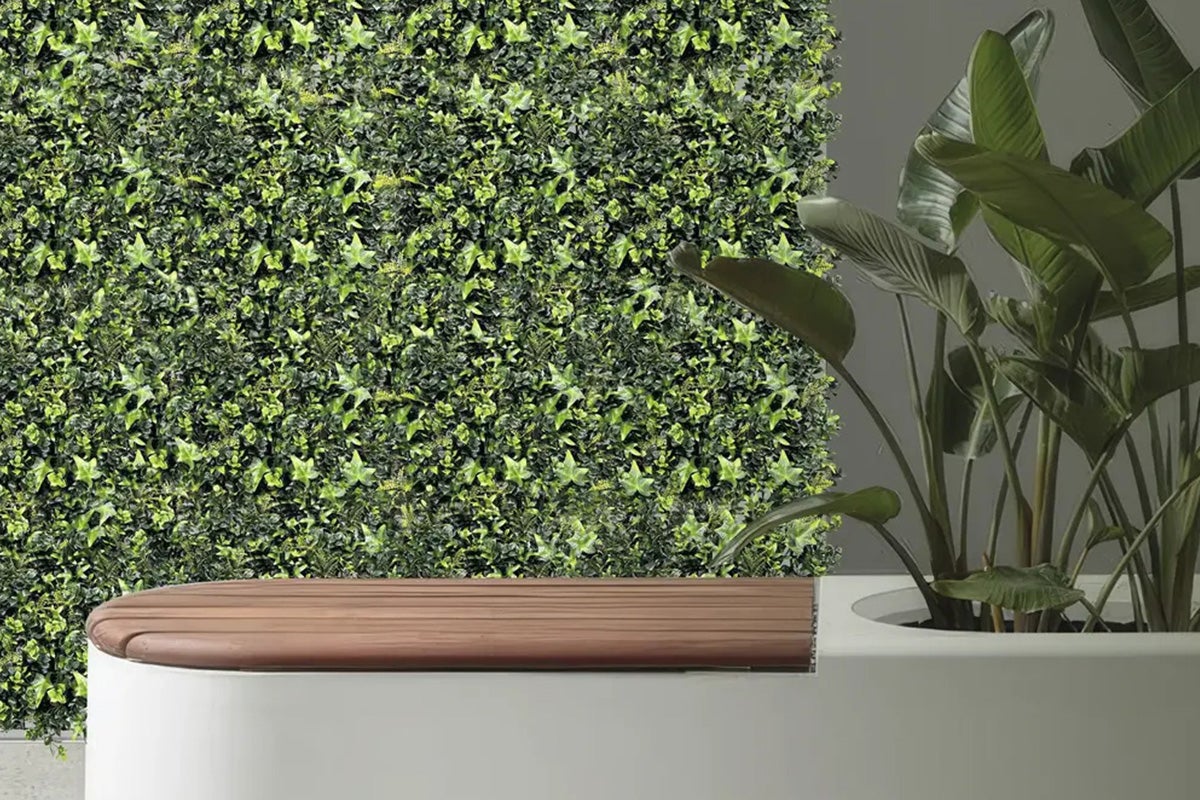 Perete decorativ cu iederă artificială, bancă cu blat din lemn și plantă de interior într-un ghiveci.