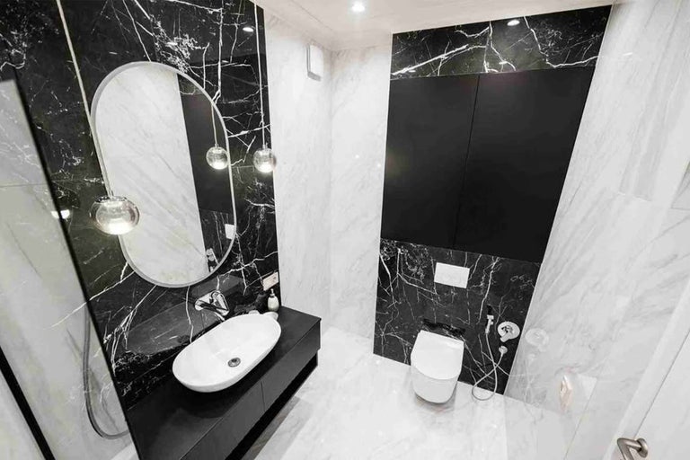 Baie modernă cu plăci cu aspect de marmură, lavoar alb pe un mobilier negru, oglindă ovală și vas WC suspendat.