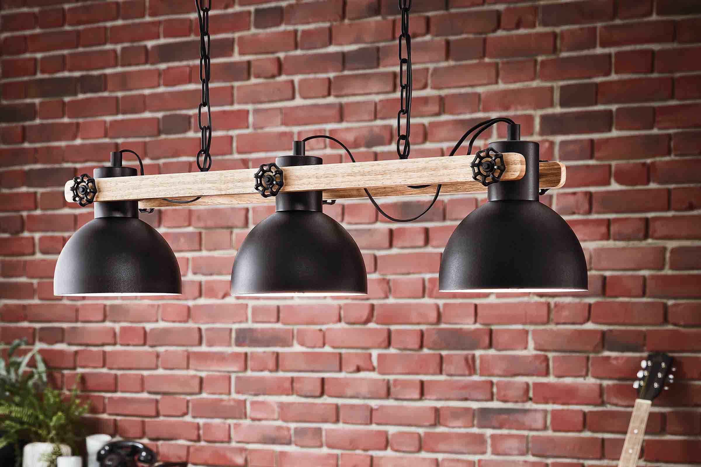 Lampa suspendată cu trei abajururi negre și bară de lemn în fața unui perete de cărămidă