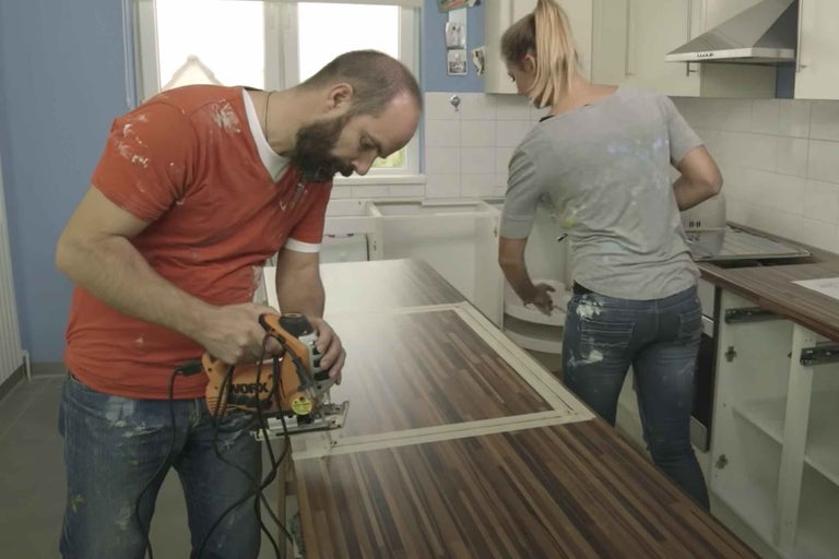 Două persoane renovează o bucătărie: O persoană taie un blat de lucru cu un ferăstrău vertical, cealaltă persoană instalează un dulap inferior.