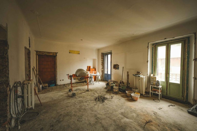 Interior luminos în timpul unei renovări, cu o betonieră, găleți și materiale de construcție pe o podea de beton brut.