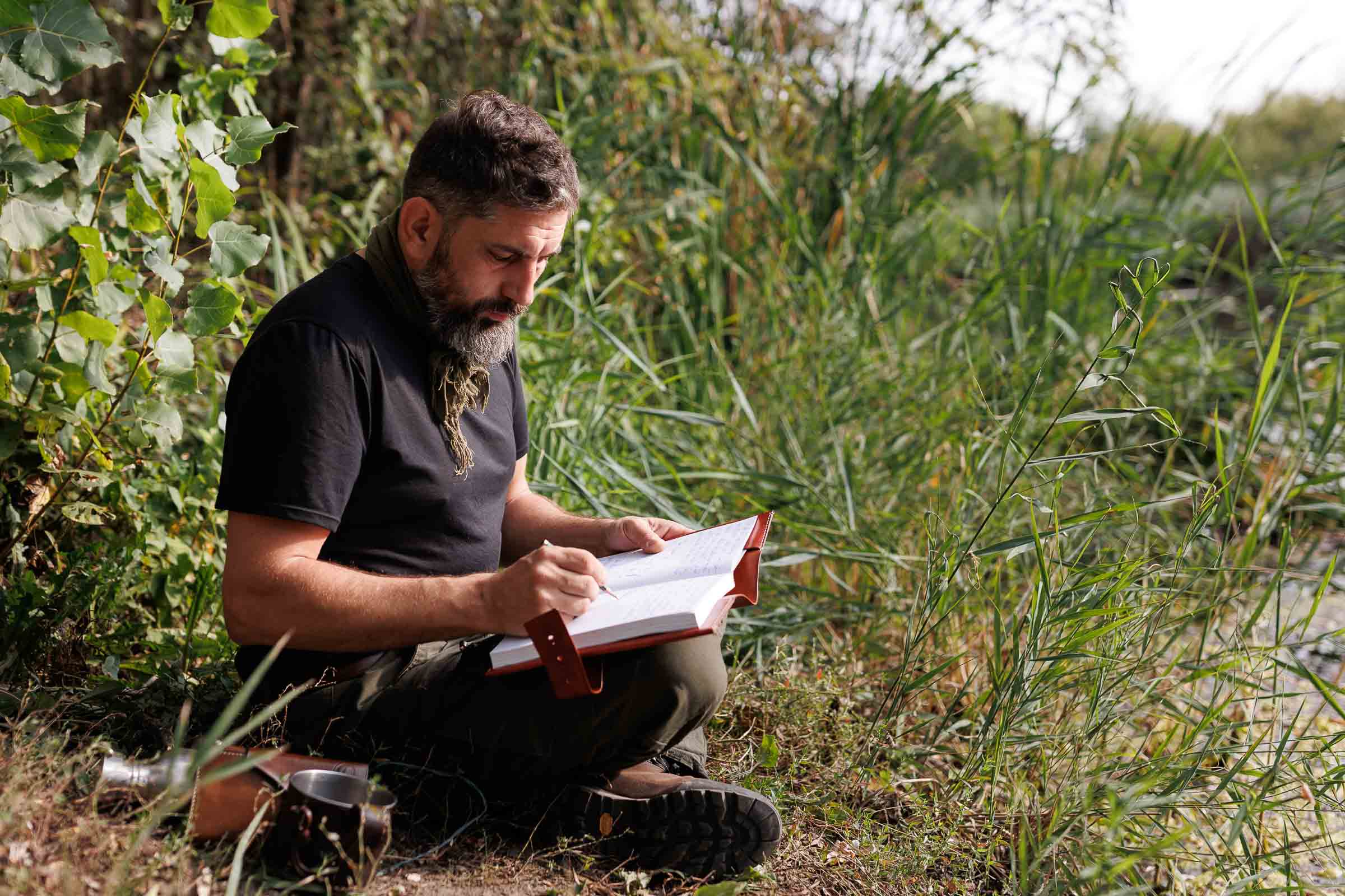 Un bărbat stă așezat în natură și scrie într-un caiet. În fundal se văd plante.