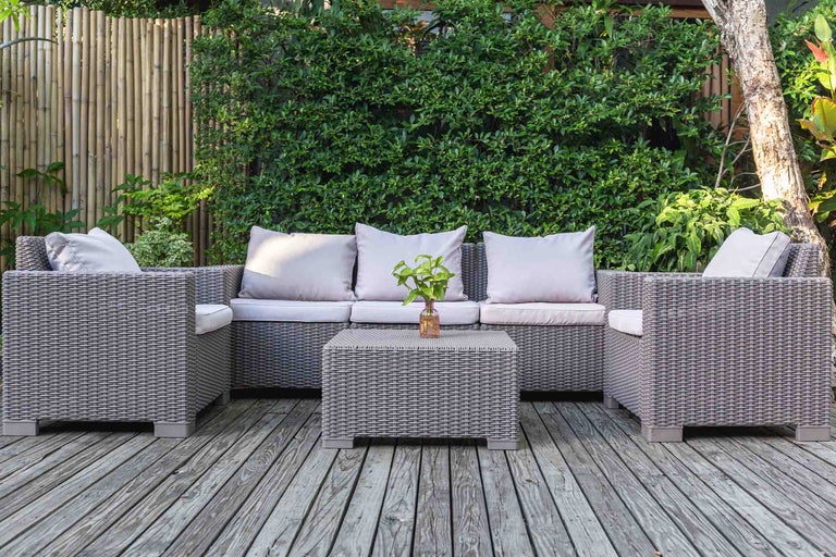 Set de mobilier de grădină din ratan sintetic gri cu canapea, două fotolii și masă pe o terasă din lemn în fața unui gard viu.