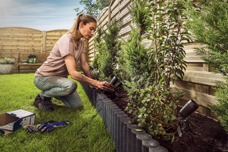 Femeie instalează iluminat de grădină într-un pat de flori cu plante și borduri de gazon