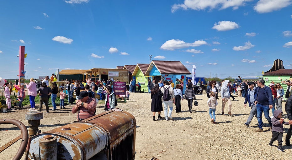 O scenă animată cu oameni plimbându-se într-o piață în fața unor căsuțe și a unui tractor vechi.