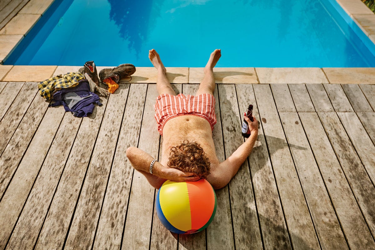 Bărbat care se relaxează pe o terasă din lemn lângă o piscină cu apă albastră, cu capul sprijinit pe o minge de plajă colorată.