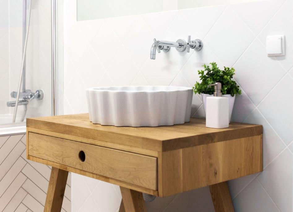 Baie luminoasă cu un dulap de lavoar din lemn, un lavoar pe blat alb cu model ondulat și o baterie de perete pe fundal de plăci ceramice tip romb.