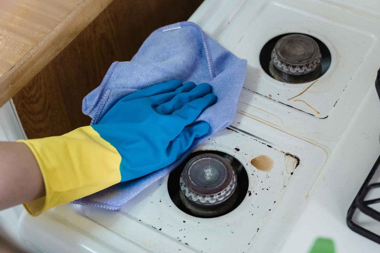 Curățarea unui aragaz murdar cu o cârpă albastră din microfibră și mănuși de protecție