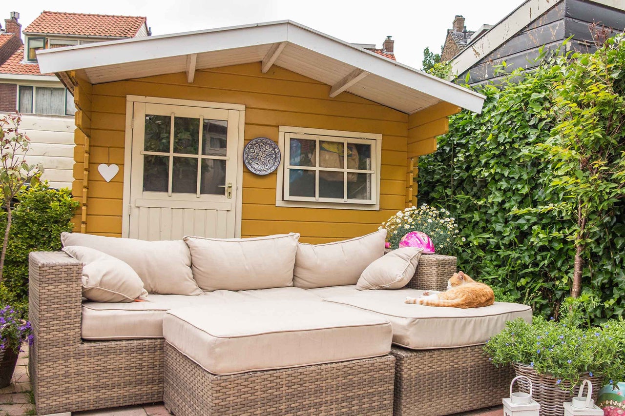 Scenă cu căsuță de grădină galbenă și canapea din ratan cu perne