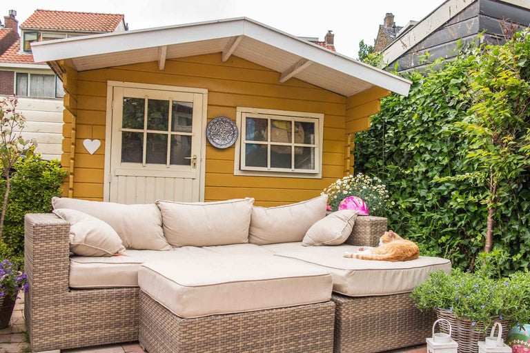 Scenă cu căsuță de grădină galbenă și canapea din ratan cu perne