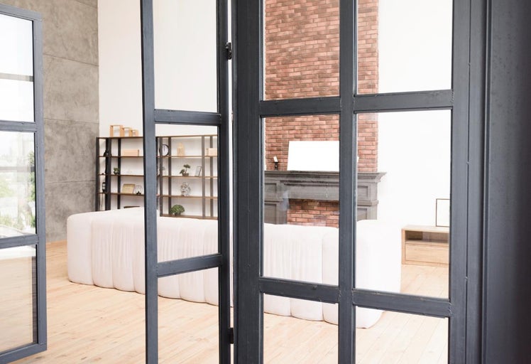 Vedere printr-o ușă deschisă spre o sufragerie luminoasă cu o canapea albă, o bibliotecă și un șemineu