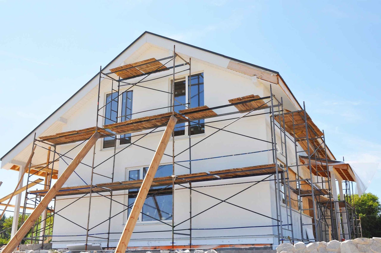 Casă unifamilială în construcție cu schelă și scânduri de lemn în fața fațadei deschise la culoare.