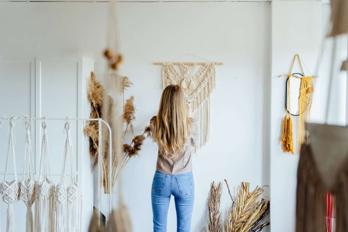 Femeie aranjând decorațiuni de perete macrame într-o cameră luminoasă cu flori uscate și oglindă