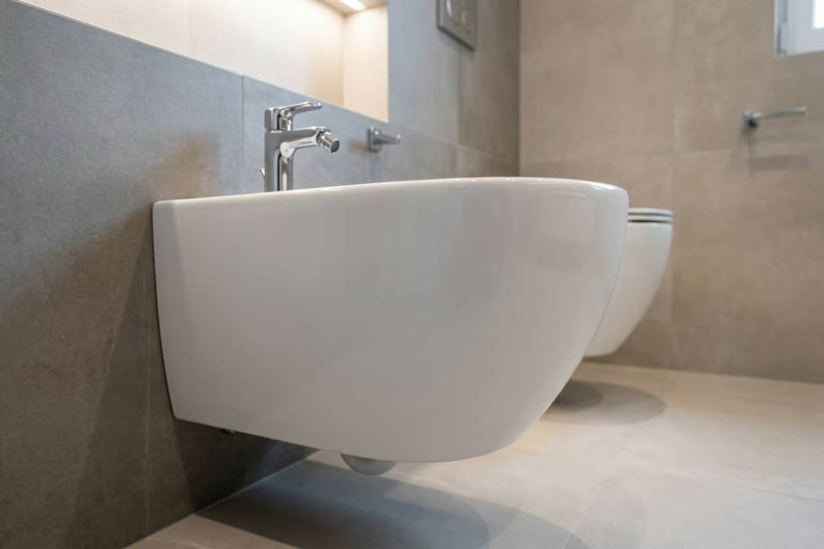 Bideu și toaletă suspendate din ceramică albă într-o baie modernă cu gresie gri și baterie cromată.