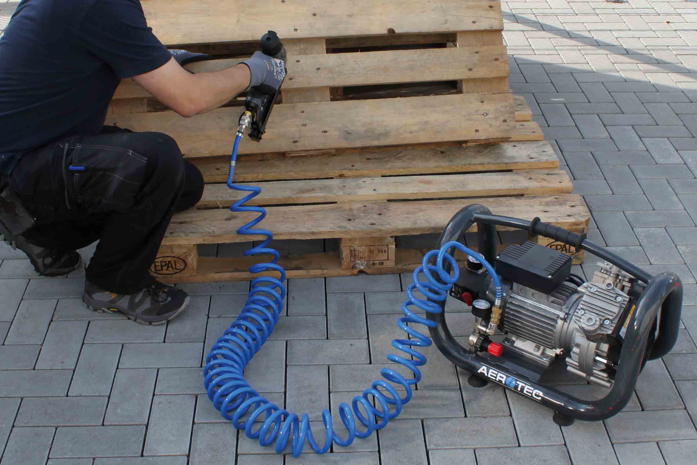 O persoană fixează un palet din lemn cu un ciocan pneumatic, conectat la un compresor Aerotec printr-un furtun albastru