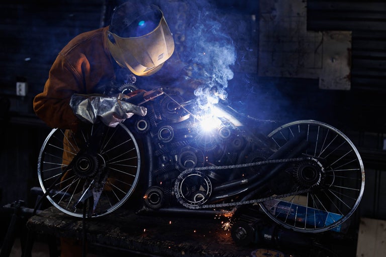 Sudor cu cască de sudură și mănuși lucrând la o sculptură din roți metalice și roți dințate.