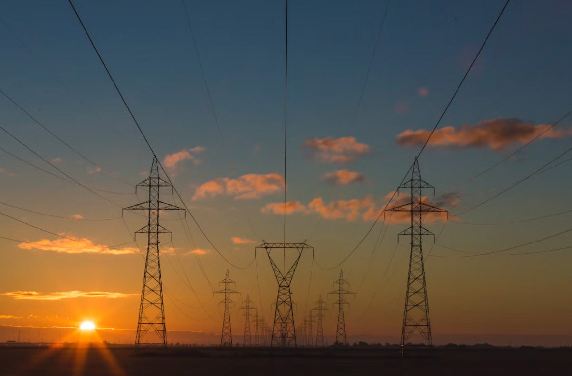 Stâlpi de înaltă tensiune cu linii electrice aliniați în timpul unui apus de soare.