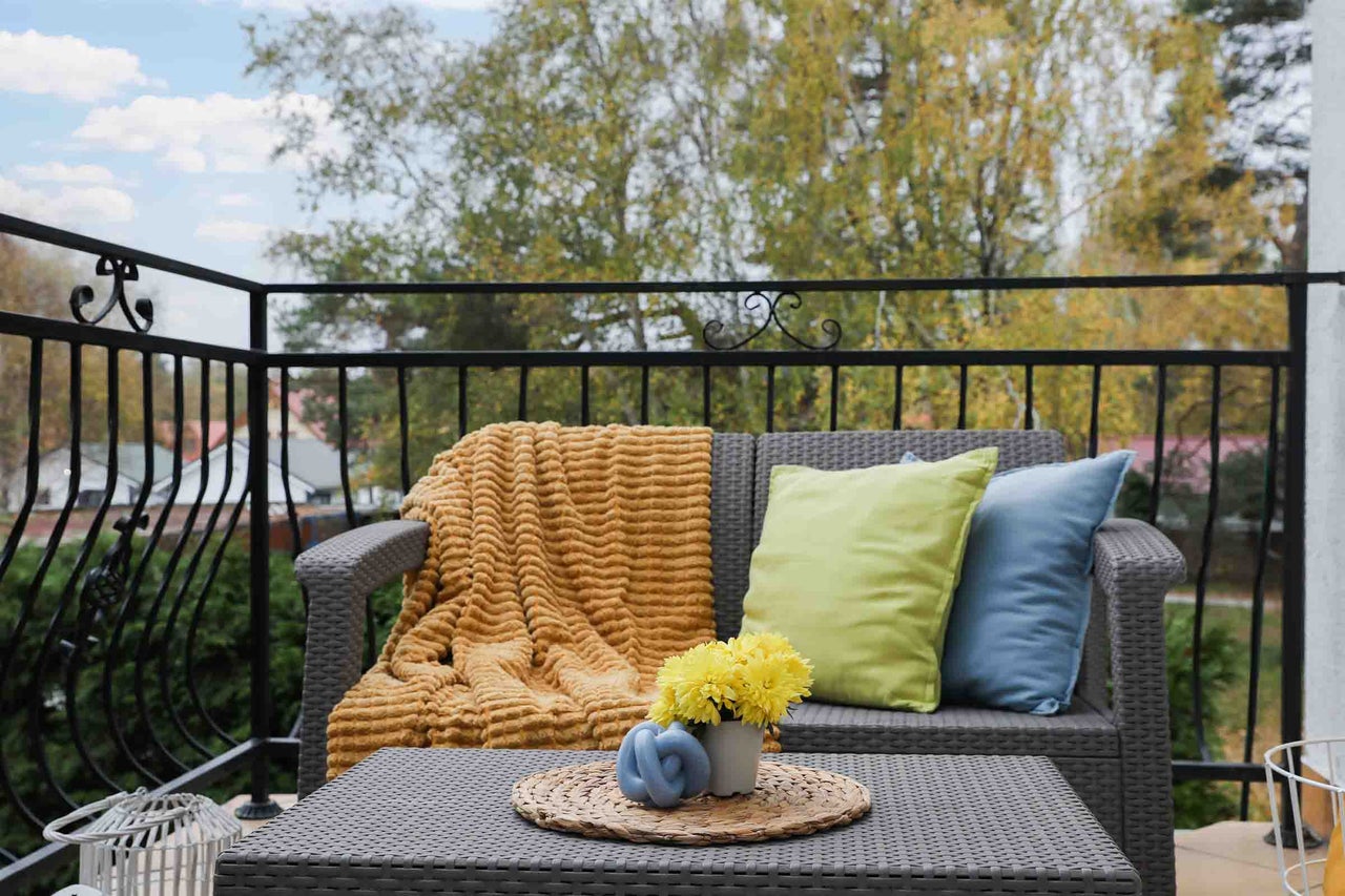 Canapea de grădină cu două locuri din ratan gri pe un balcon, cu pătură galbenă, perne și măsuță de cafea, pe fundalul unor copaci de toamnă.