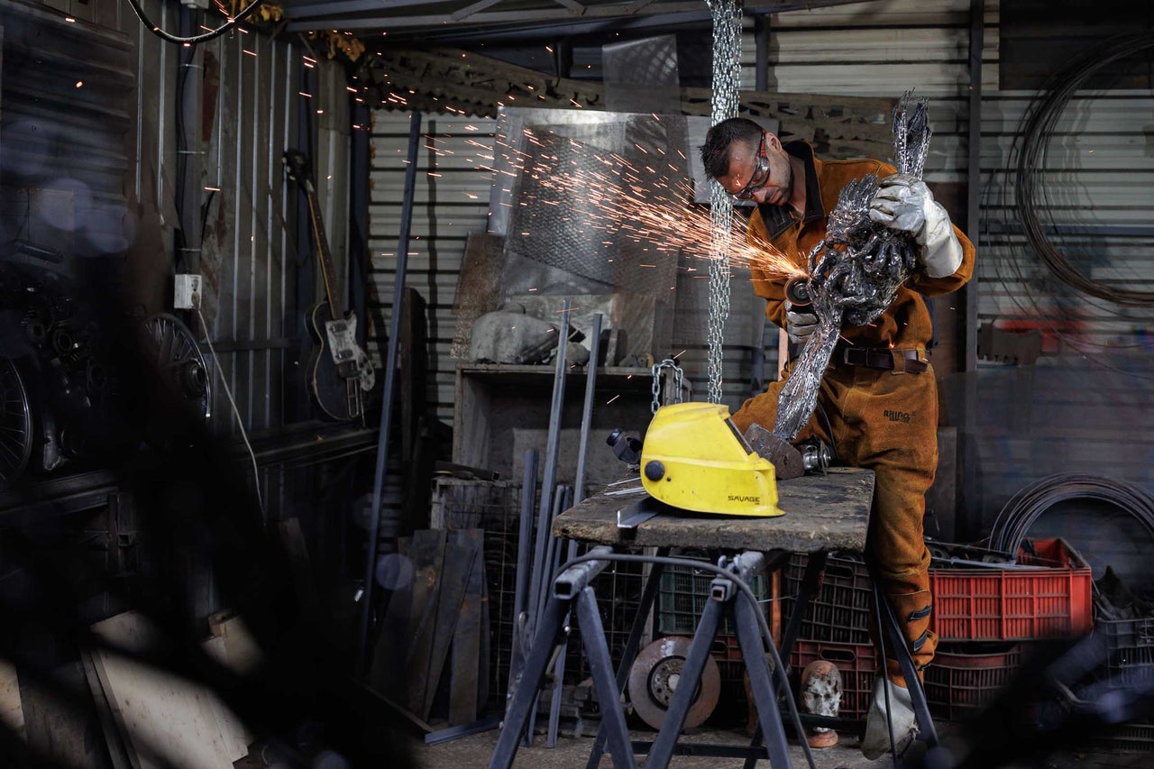 Un bărbat în echipament de protecție prelucrează o sculptură metalică cu un polizor unghiular într-un atelier.