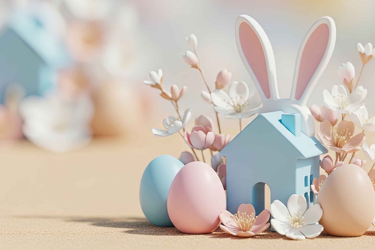 Decorațiune de Paște luminoasă cu o căsuță albastră, urechi de iepuraș, ouă de Paște colorate și flori de primăvară într-un aranjament ordonat.