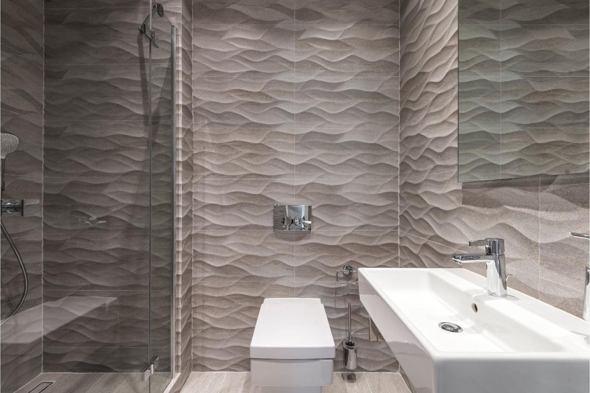 Baie modernă cu duș, toaletă și lavoar în fața unui perete cu plăci ceramice cu aspect ondulat